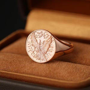 May include: A rose gold signet ring featuring a detailed phoenix design. The ring has a polished finish and is set against a brown velvet background. The phoenix is depicted with outstretched wings within a circular frame.