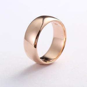 Puede incluir: Un anillo de oro rosa pulido con una superficie lisa y abombada y un perfil redondeado. El metal tiene un tono rosa metálico cálido con propiedades reflectantes, mostrando un diseño de banda sólida y simple sin grabados ni engastes de piedras.