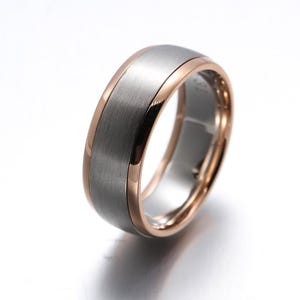 May include: A band ring featuring a wide, brushed silver-tone centre inlay flanked by polished rose gold-tone edges. The ring has a smooth, rounded profile with a metallic finish, showcasing a distinct contrast between the matte grey centre and the shiny, warm-toned metallic borders.