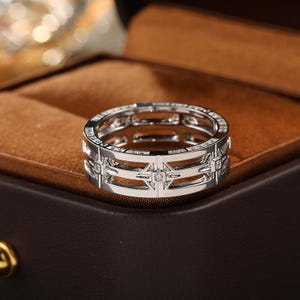 Può includere: Un anello d'argento con un design geometrico. L'anello presenta una serie di ritagli rettangolari e quadrati, con piccole gemme chiare incastonate nel design. L'anello è esposto in un portagioie marrone.