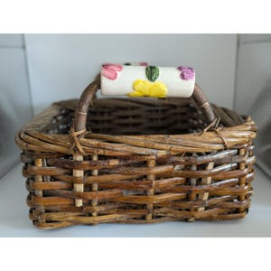 May include: A rectangular brown woven wicker basket featuring a sturdy handle. The handle is accented with a white ceramic grip decorated with hand-painted pink, yellow, and purple floral motifs and green leaves, integrated into the basket frame.