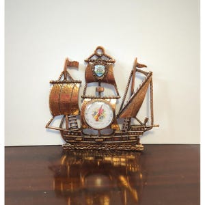 May include: A copper-toned metal wall hanging shaped like a sailing ship with three masts. The central mast features a round thermometer dial with a floral graphic and numerical markings. The ship has textured sails and a small shield emblem.