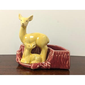 May include: A ceramic planter featuring a standing yellow deer and a smaller fawn resting together. The deer figures are positioned in front of a textured, reddish-brown log-shaped base. The piece has a glossy finish and a hollow interior section.