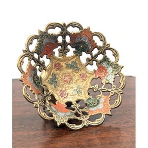May include: A round brass bowl featuring an intricate openwork filigree border and a central basin decorated with embossed floral patterns. The metal surface is accented with enamel details in shades of orange, green, and white throughout the design.