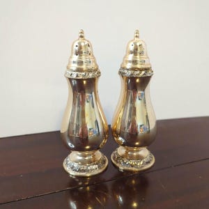 May include: A pair of polished gold-tone metal salt and pepper shakers featuring a curved, hourglass-like body, a rounded top with small dispensing holes, and a decorative textured band around the base and the neck of each shaker.
