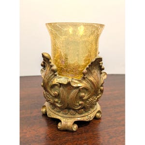 May include: A votive candle holder featuring a cylindrical yellow crackle glass insert held within an ornate, antique gold-toned metal base. The base displays intricate scrollwork, leaf-like motifs, and a scalloped edge, supported by small feet.
