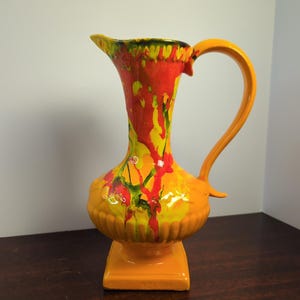 May include: A vibrant, mid-century modern ceramic pitcher. The pitcher features a bright orange base and handle, with a tall, flared body adorned with dripped red, yellow, and green paint. The pitcher stands on a square base.