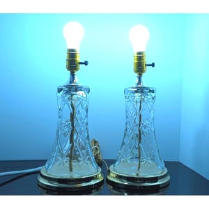 May include: A pair of table lamps featuring clear cut glass bases with a diamond and star pattern. Each lamp has a gold-toned metal socket, a round gold-toned base, and a single glowing light bulb. The lamps are connected by a white power cord.