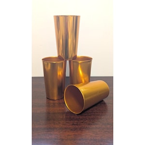 May include: A set of four metallic gold-colored tumblers with a smooth, reflective finish. Three of the tumblers are standing upright in a pyramid formation, while the fourth tumbler is positioned horizontally in the foreground.