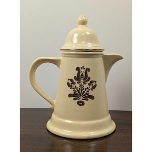 May include: A cream-colored ceramic coffee pot featuring a brown floral pattern on the front. The pot has a curved handle, a pointed spout, and a matching lid with a small knob handle on top. The design is symmetrical and centered on the body.