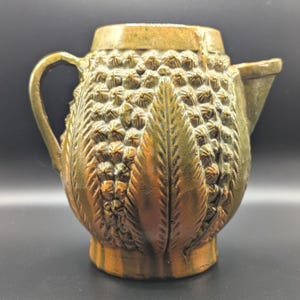 May include: A bronze-colored decorative pitcher with a textured pineapple design. The pitcher has a handle and a spout, and the surface is detailed with a pattern of overlapping scales and leaves. The overall shape is rounded, with a slightly flared base.