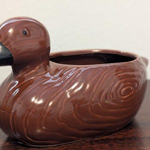 May include: A brown ceramic duck-shaped planter with a glossy finish. The duck has a black beak and a detailed wood-grain texture. The planter has a wide opening for plants or decorative items.