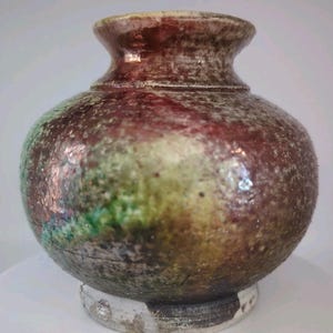 May include: A handcrafted ceramic vase with a bulbous body and a narrow neck. The vase features a mottled glaze in shades of brown, red, green, and gold. The base is a lighter color, and the overall texture is rough.