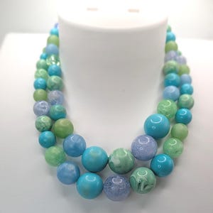 May include: A two-strand necklace featuring large, round beads in shades of blue, green, and lavender. The beads have a marbled or speckled appearance, creating a unique texture. The necklace is displayed on a white bust.