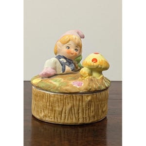 May include: A ceramic trinket box featuring a sculpted lid with a blonde elf wearing a pink hat and blue outfit, resting beside a yellow mushroom with red spots. The base of the box is textured to resemble a brown tree stump with vertical bark details.