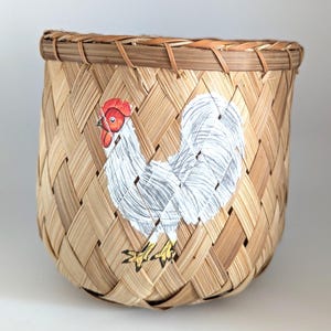 May include: Woven bamboo basket with a hand-painted rooster design. The rooster is white with a red comb and yellow feet. The basket is a natural tan color with a woven pattern.