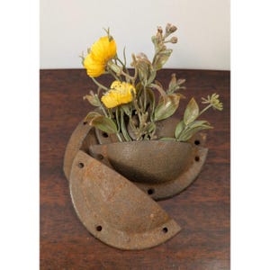 May include: A collection of three semi-circular, cast iron cup pulls with a weathered, rusted brown finish. Each pull features two screw holes on the flat mounting flange. A small bouquet of artificial yellow flowers is tucked into the pulls.