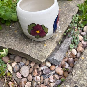 May include: A hand-painted ceramic plant pot in off-white, featuring a burgundy flower with a yellow centre and green leaves. The pot is cylindrical and rests on a stone surface. The design is likely hand-painted.