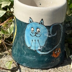 May include: A hand-painted ceramic mug with a cat motif. The mug has a light blue cat illustration on a dark teal base, with a cream-coloured top. An orange owl is also painted on the mug.