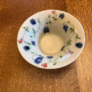 May include: A small, round, off-white ceramic bowl with a wide, sloped rim. The interior and rim are decorated with a hand-painted pattern of delicate green vines, small blue flowers, and tiny pink heart-shaped blossoms scattered across the surface.