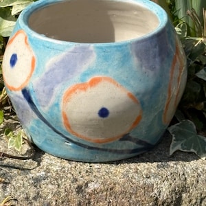 May include: A round, hand-painted ceramic planter with a light blue background and floral designs in orange, white, and blue. The planter is sitting on a stone surface and surrounded by greenery.