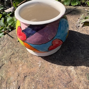 May include: A round ceramic planter featuring a hand-painted design of abstract shapes and hearts in vibrant colours including blue, purple, red, orange, pink, and lime green. The pot has a smooth white interior and a matching white circular base.