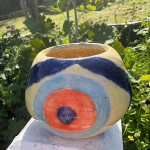 Op de afbeelding: Een handbeschilderde keramische kom met een uniek ontwerp. De kom heeft een gele basis met een donkerblauwe band, lichtblauwe en oranje concentrische cirkels. De binnenkant is warmbruin van kleur. De kom staat buiten met groen op de achtergrond.