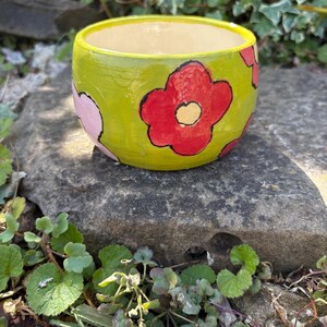 May include: A small, round ceramic bowl with a glossy lime green exterior featuring hand-painted red and light pink flower motifs outlined in black. The interior of the bowl is a smooth, solid cream colour with a wide, open circular rim.