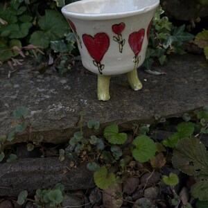 May include: A white ceramic planter with a wide, flared rim and three short, pale green legs. The exterior surface is decorated with hand-painted red heart motifs, each featuring a thin black stem and leaf detail, arranged around the bowl.