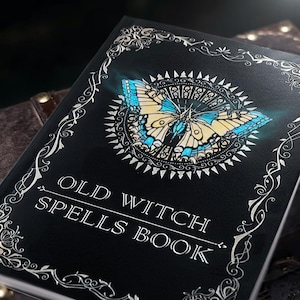 Puede incluir: Un libro negro titulado "OLD WITCH SPELLS BOOK" con un dise&ntilde;o detallado de mariposa en azul, amarillo y blanco. La portada tiene bordes y detalles decorativos plateados, creando una est&eacute;tica m&iacute;stica.