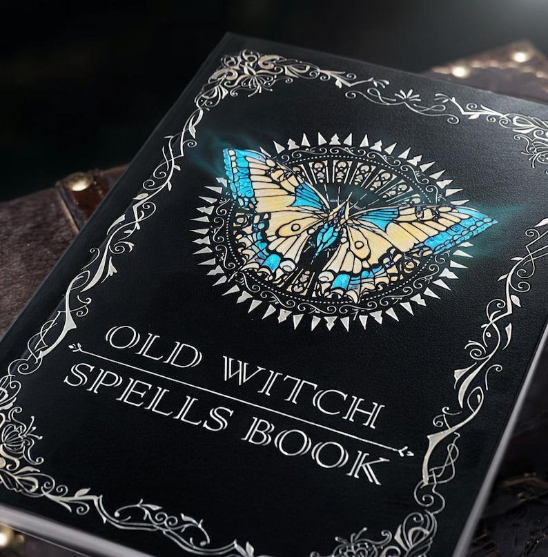 Puede incluir: Un libro negro titulado "OLD WITCH SPELLS BOOK" con un dise&ntilde;o detallado de mariposa en azul, amarillo y blanco. La portada tiene bordes y detalles decorativos plateados, creando una est&eacute;tica m&iacute;stica.