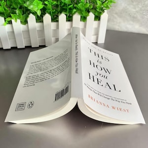 May include: An open book titled "This Is How You Heal" by Brianna Wiest. The cover and pages are white with black and red text. The book is from Thought Catalog Books.
