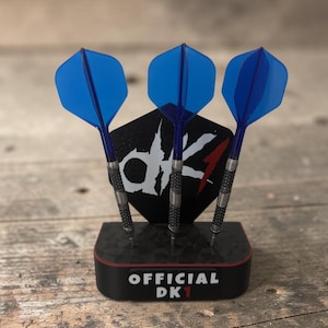 Könnte beinhalten: Drei Darts mit blauen, durchscheinenden Flights und strukturierten silbernen Barrels stecken in einem schwarzen Ständer. Der Ständer hat eine sechseckige Rückwand mit dem weißen Text "dk1" und eine Basis mit dem Text "OFFICIAL DK1".