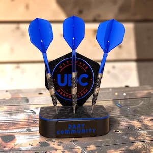 Könnte beinhalten: Ein Set aus drei Darts mit leuchtend blauen Flights und texturierten schwarzen Barrels, die aufrecht in einem schwarzen Ständer stehen. Der Ständer hat eine sechseckige Rückplatte mit dem Text "ULTIMATE DART COMMUNITY" und "UDC" in Blau.