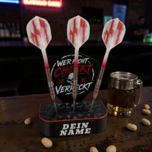 Könnte beinhalten: Drei Dartpfeile mit rot-weiß gemusterten Flights werden in einem schwarz-roten Halter präsentiert. Der Halter trägt den Text "DEIN NAME" auf der Vorderseite. Ein Bierkrug und Erdnüsse sind ebenfalls zu sehen.