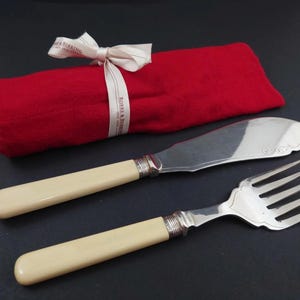 Op de afbeelding: Een botermes en vork set met ivoorkleurige handvatten en zilverkleurige bladen. Naast de set ligt een opgerolde rode doek, vastgebonden met een lint met de tekst "ROBBS & BERKING".