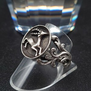 May include: A silver ring featuring a raised oval emblem with a detailed depiction of a deer. The ring has an ornate, scrolling design on the band. The ring is displayed on a clear, angled stand.