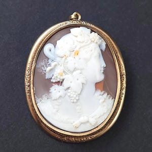 Pode incluir: Pingente de camafeu oval com moldura dourada. O camafeu apresenta um perfil branco de mulher com folhas de uva e cachos de uva. O fundo é marrom quente. O pingente está sobre um fundo escuro.