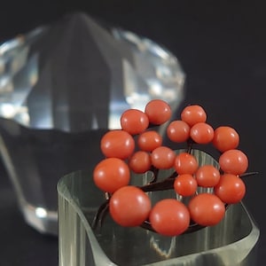 Puede incluir: Broche de coral vintage con un grupo de cuentas redondas de coral naranja-rojo. El broche está engarzado sobre una base de metal oscuro y se exhibe sobre un soporte de vidrio transparente. Un gran cristal facetado en el fondo.