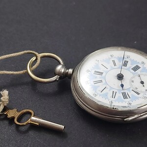 Könnte beinhalten: Eine silberne Taschenuhr mit weißem Zifferblatt, blauen Akzenten und schwarzen römischen Ziffern. Die Uhr hat ein kleines Sekunden-Zifferblatt und einen goldenen Schlüssel, der an einer Schnur befestigt ist. Ein klassischer Zeitmesser.