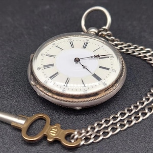 Op de afbeelding: Een zilveren zakhorloge met een witte wijzerplaat, zwarte Romeinse cijfers en een zilveren ketting. Het horloge heeft een sleutel. Het horloge ligt op een donkere ondergrond.