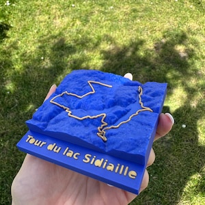 May include: A blue 3D model of a lake with a gold outline and text that reads "Tour du lac Sidiaille". The model is held in a hand against a background of green grass.