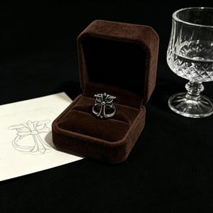 May include: A silver ring with a decorative cross design is displayed inside a brown velvet ring box. A sketch of the ring is on a white card. A clear glass with water is in the background.