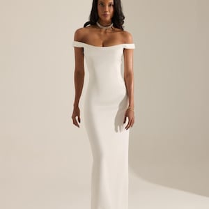 Könnte beinhalten: Ein bodenlanges, schulterfreies weißes Kleid. Das Kleid hat ein eng anliegendes Oberteil und einen fließenden Rock. Das Model trägt eine zarte Halskette und ein Armband. Der Hintergrund ist einfarbig weiß.