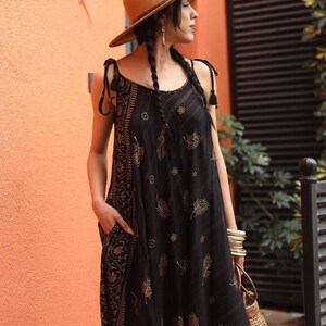 May include: A black patterned dress with a brown hat. The dress has a floral design and pockets. The hat is brown with a dark brown band. The dress has tie straps and a loose fit. The person is wearing bracelets and carrying a small woven bag.