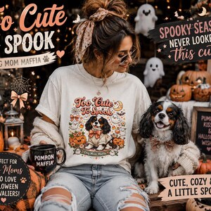 Op de afbeelding: Een crèmekleurig t-shirt met een afbeelding van een Cavalier King Charles Spaniel met een vlinderdas, omringd door pompoenen, spoken en sterren, met de tekst Too Cute To Spook en Dog Mom Maffia op de voorzijde van het kledingstuk.