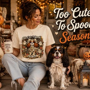 Op de afbeelding: Een crèmekleurig t-shirt met een afbeelding van een Cavalier King Charles Spaniel verkleed als spook, omringd door kleine spookjes, bloemen en de tekst Too Cute To Spook Dog Mom Mafia, gecombineerd met een lichtblauwe spijkerbroek.