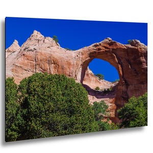 Puede incluir: Fotografía de paisaje de un arco de roca natural bajo un cielo azul claro y brillante. El arco es de color marrón rojizo, con árboles y arbustos verdes en primer plano. La imagen es una impresión sobre un fondo negro.