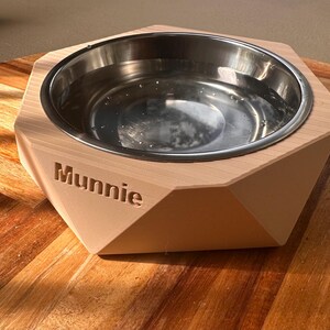 May include: A geometric wooden pet bowl with a stainless steel interior, filled with water. The name "Munnie" is engraved on the front. The bowl is on a wooden surface. The bowl is likely for a dog or cat.