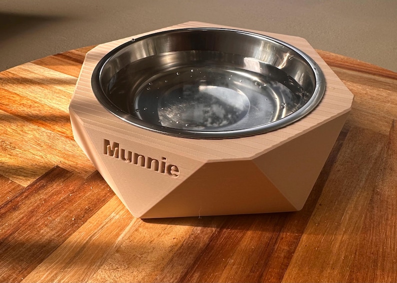 May include: A geometric wooden pet bowl with a stainless steel interior, filled with water. The name "Munnie" is engraved on the front. The bowl is on a wooden surface. The bowl is likely for a dog or cat.